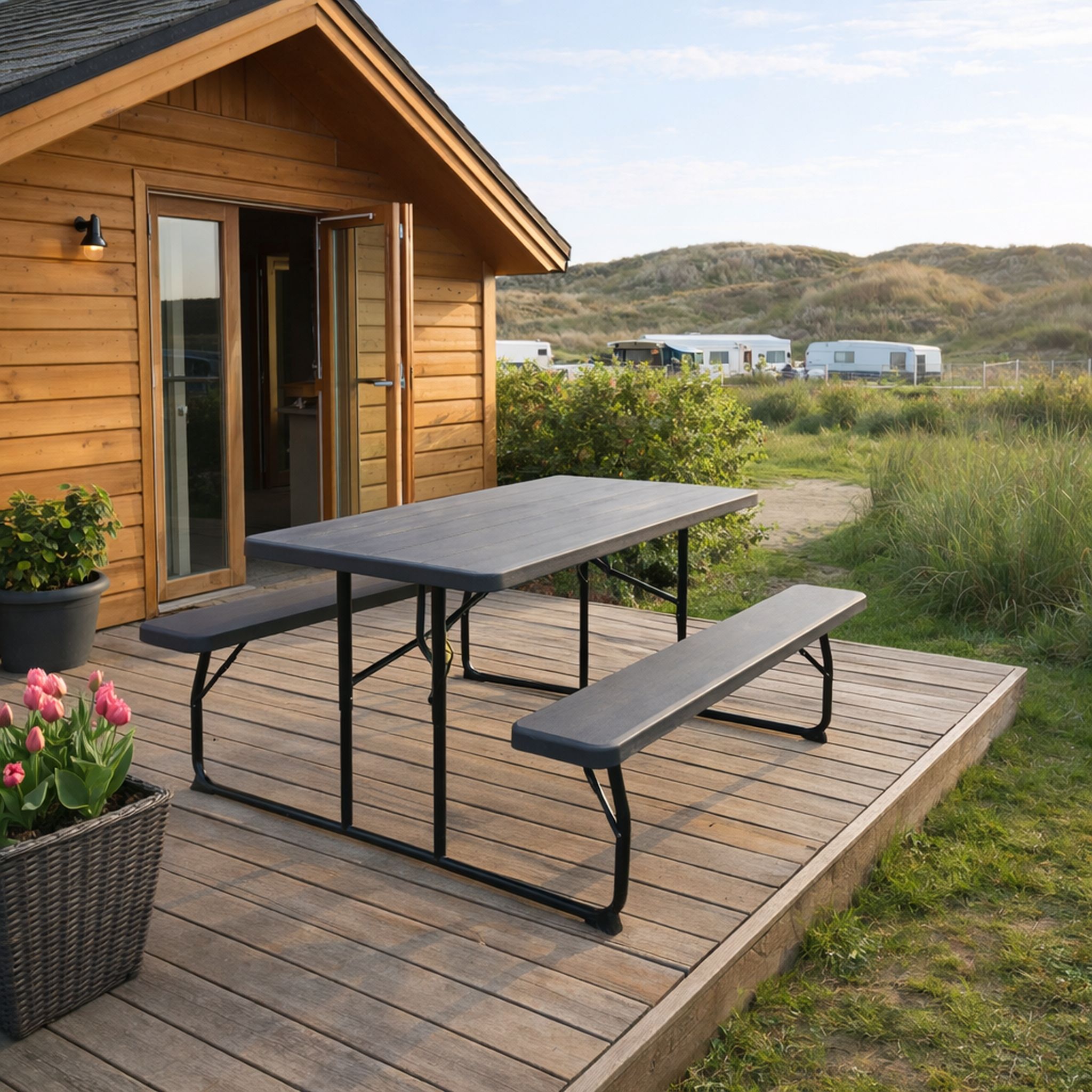 Holzhütte mit Terrasse, darauf ein Tisch mit Bänken, blühende Pflanzen daneben, Wiesen und Wohnwagen im Hintergrund.