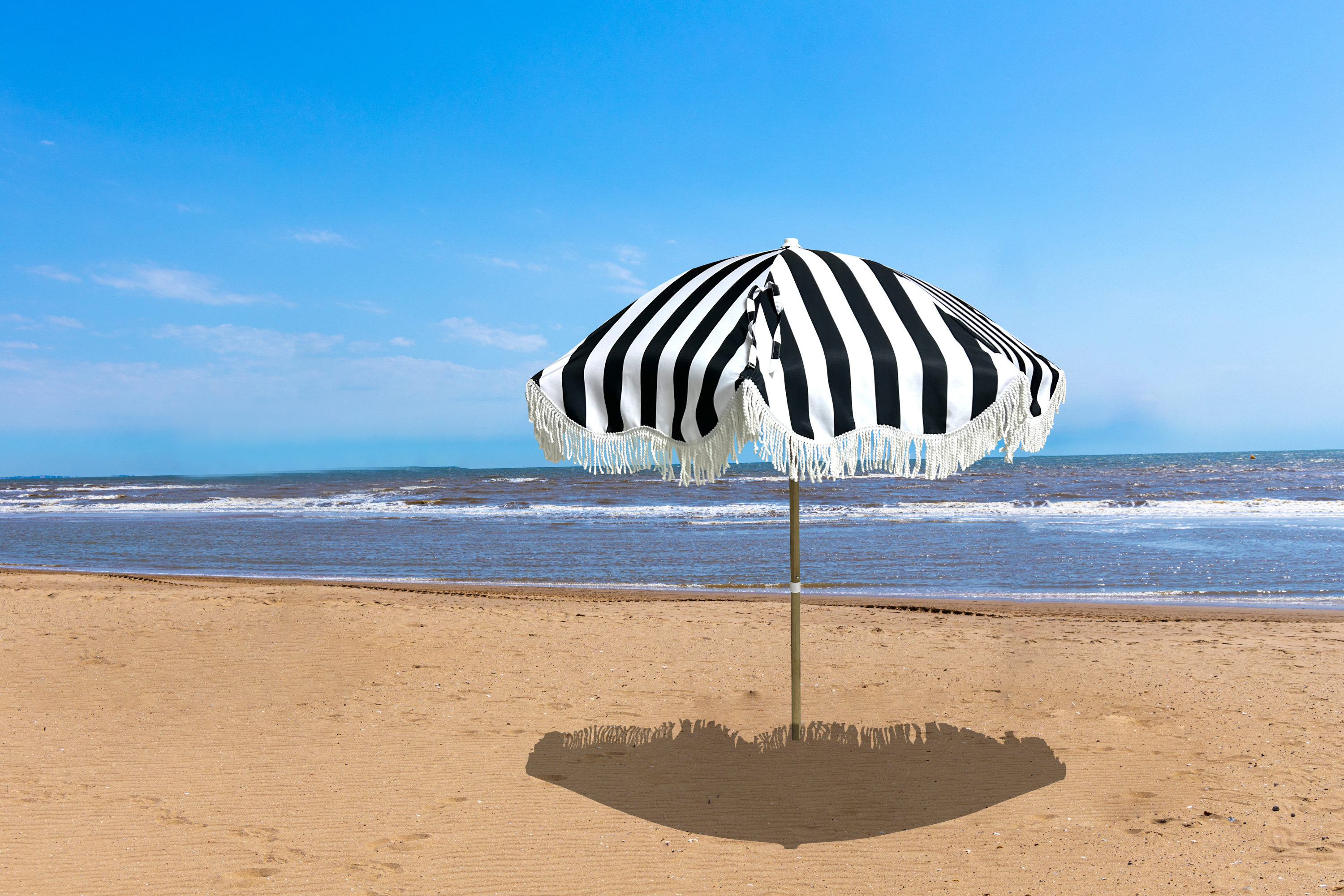Schwarz-weiß gestreifter Sonnenschirm mit Fransen steht allein am Sandstrand vor dem Meer.