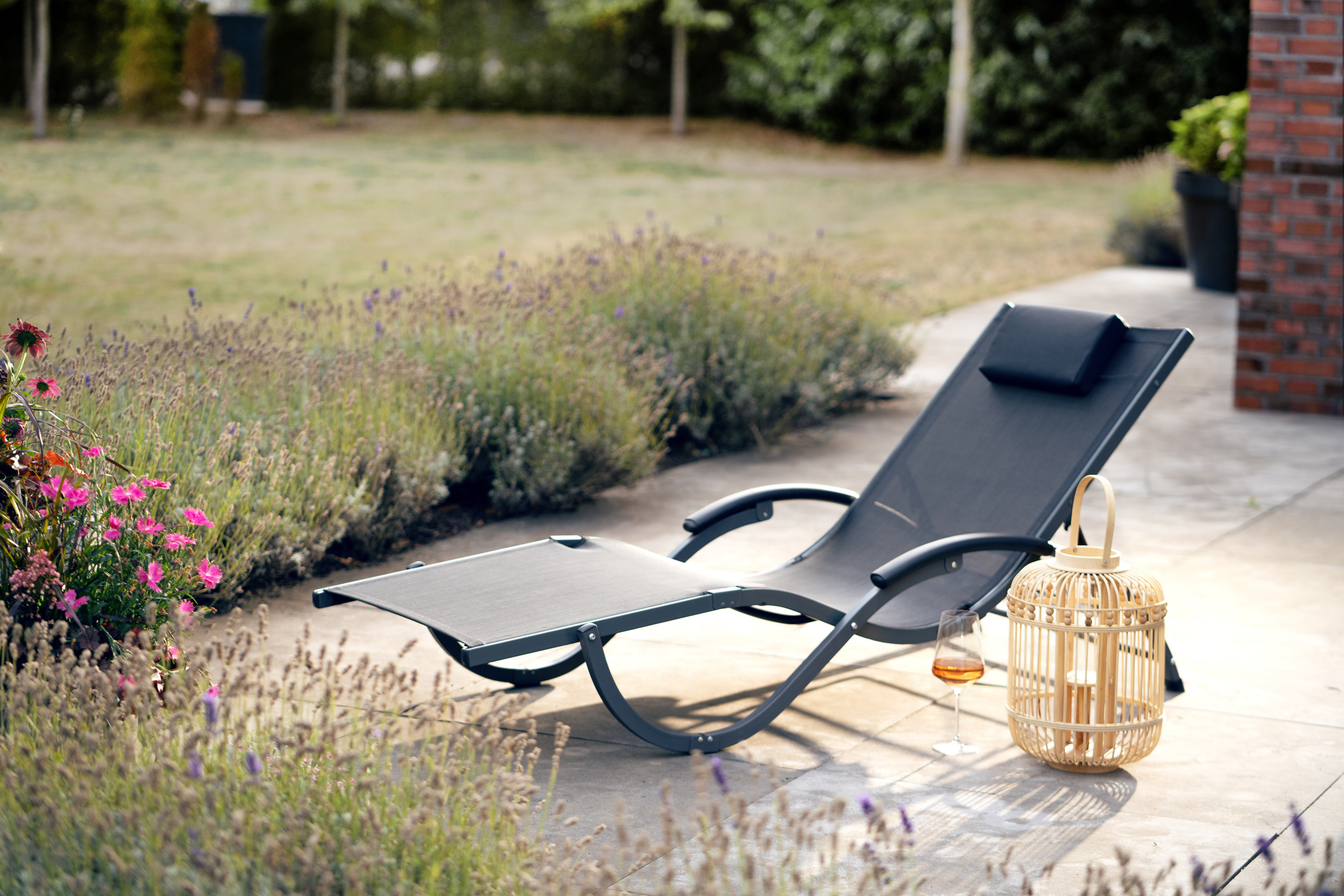 Gartenliege auf einer Terrasse, umgeben von Lavendel und anderen Blumen, mit einem Glas Getränk und einer Laterne.