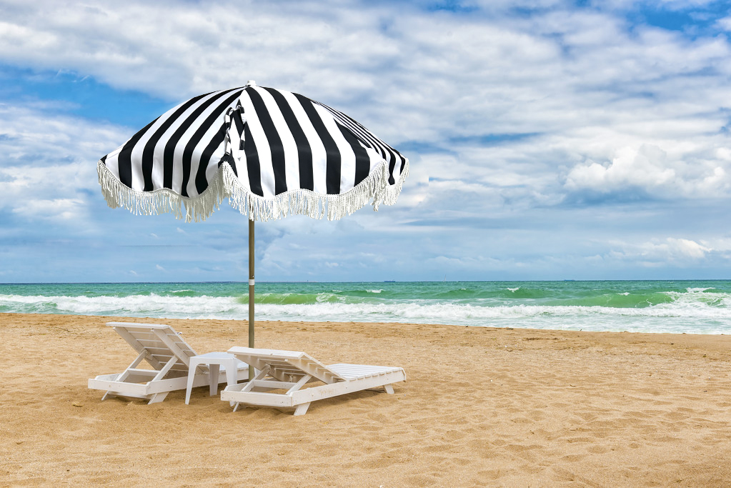 Zwei Liegestühle und ein gestreifter Sonnenschirm am Sandstrand mit Meerblick im Hintergrund.