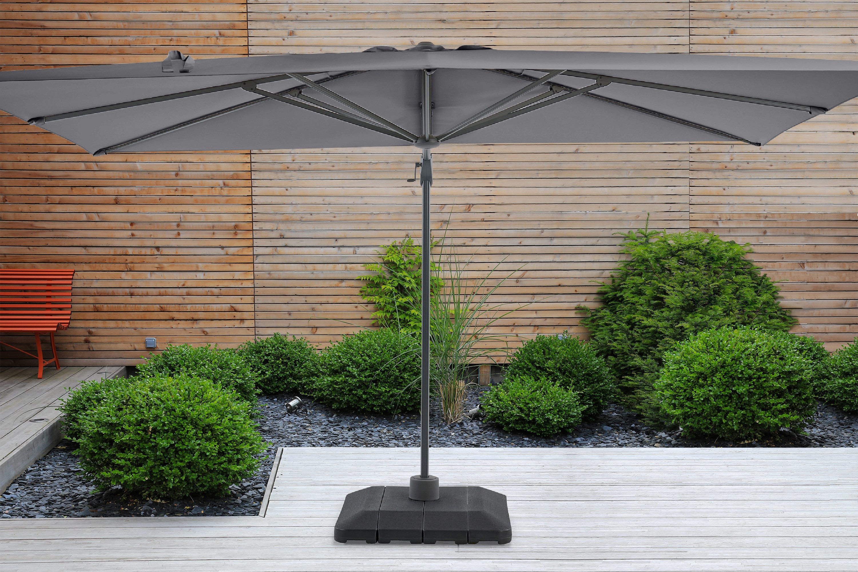 Grauer Sonnenschirm auf Terrasse mit Holzlattenwand, grünen Büschen und oranger Bank als Hintergrund.